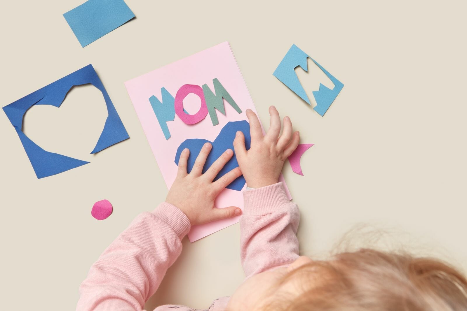 Kind 131 Close up of cute child enjoying creative process of greeting card making for mother's day. Pieces of cutted colorful paper scattered on beige background.