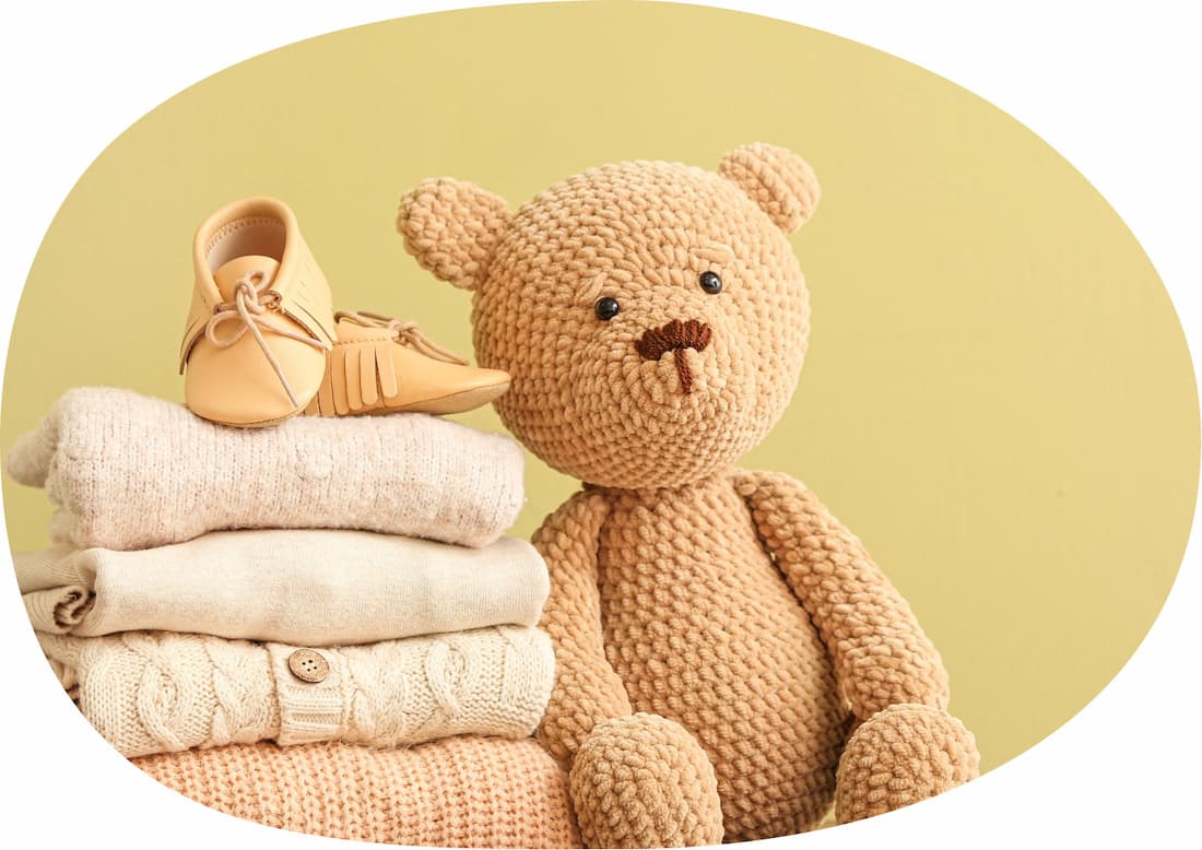 a stuffed knitted brown bear leaning against a pile of neatly folded clothing with some babies shoes on top